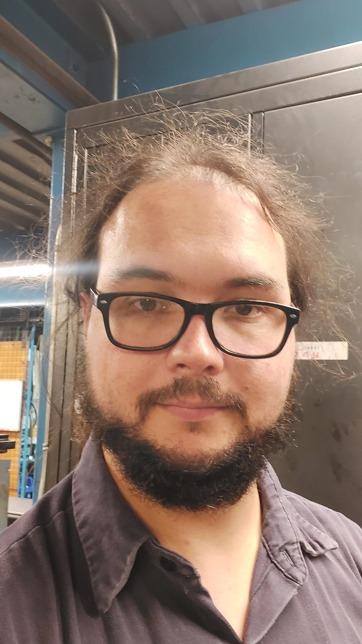 A man with tied back long black hair stands in an industrial maintenance office, photo 2